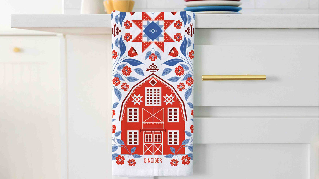 Americana Barn Tea Towel hanging from a white kitchen countertop, featuring a red folk-art barn surrounded by blue leaves, red flowers, and a patriotic quilt star motif in red, white, and blue.