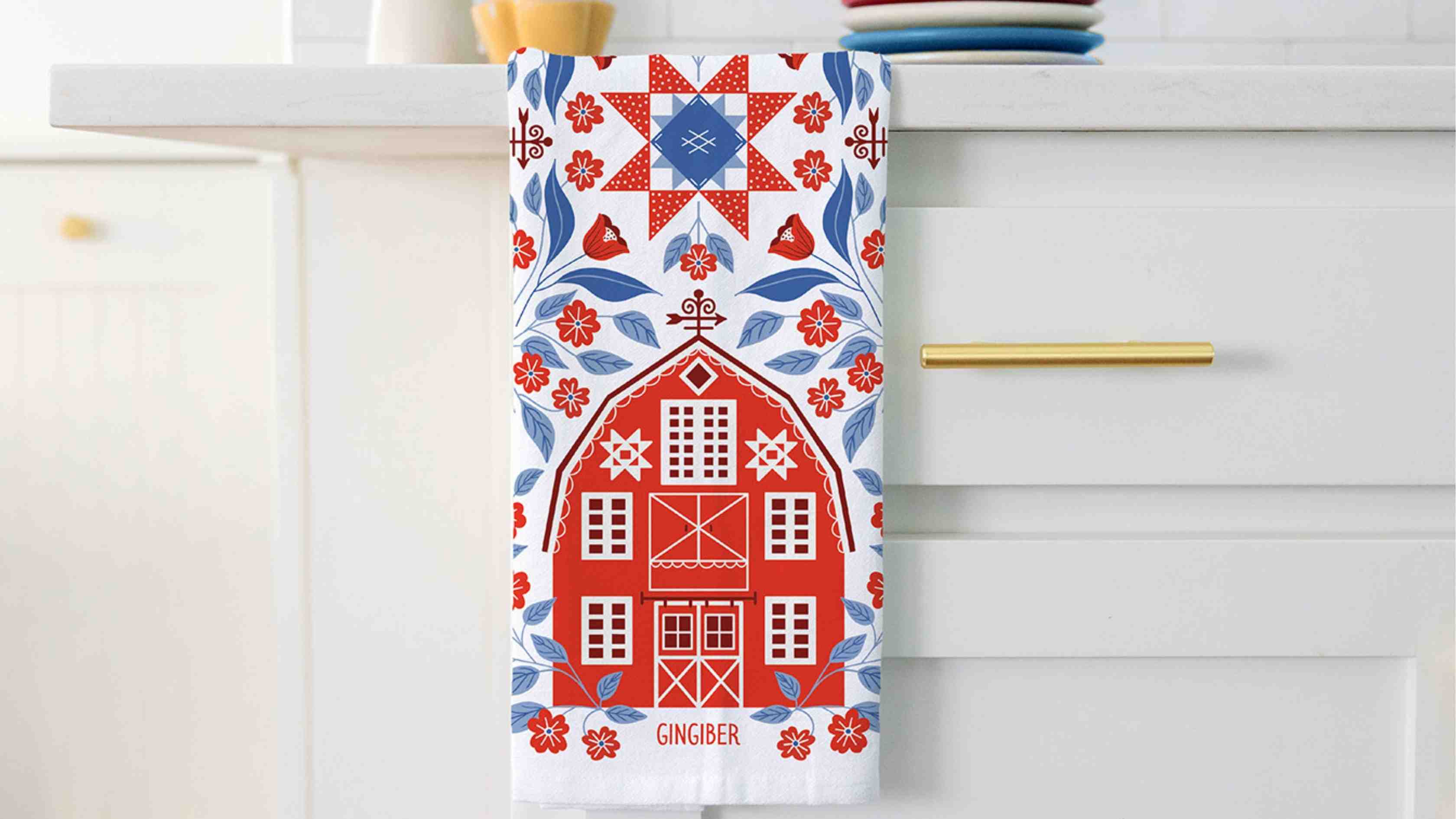 Americana Barn Tea Towel hanging from a white kitchen countertop, featuring a red folk-art barn surrounded by blue leaves, red flowers, and a patriotic quilt star motif in red, white, and blue.
