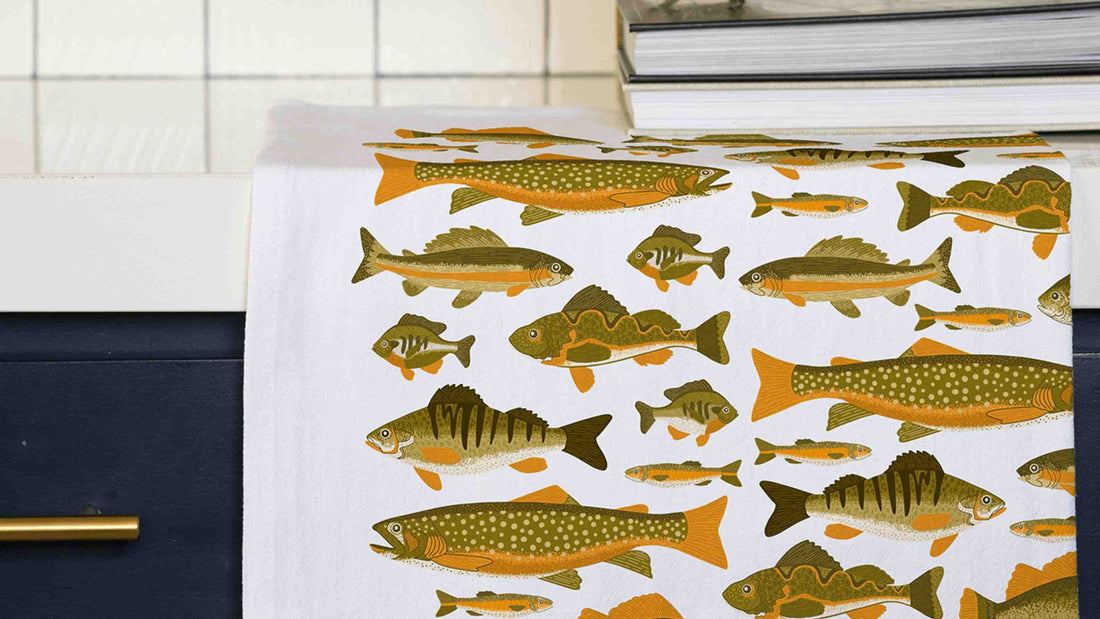 Fish Tea Towel draped over a kitchen counter, featuring detailed illustrations of freshwater fish including trout, bass, and perch in earthy green and golden orange tones.