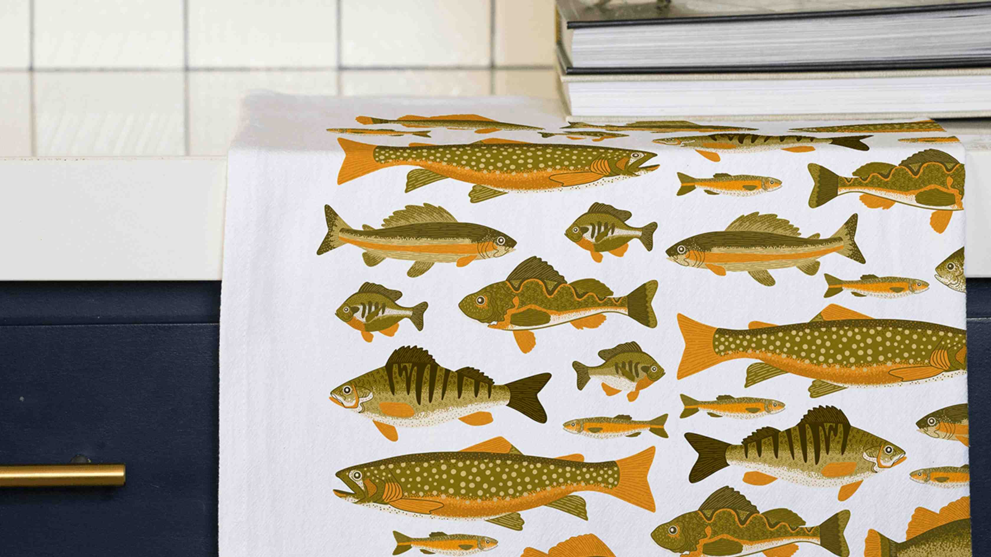 Fish Tea Towel draped over a kitchen counter, featuring detailed illustrations of freshwater fish including trout, bass, and perch in earthy green and golden orange tones.