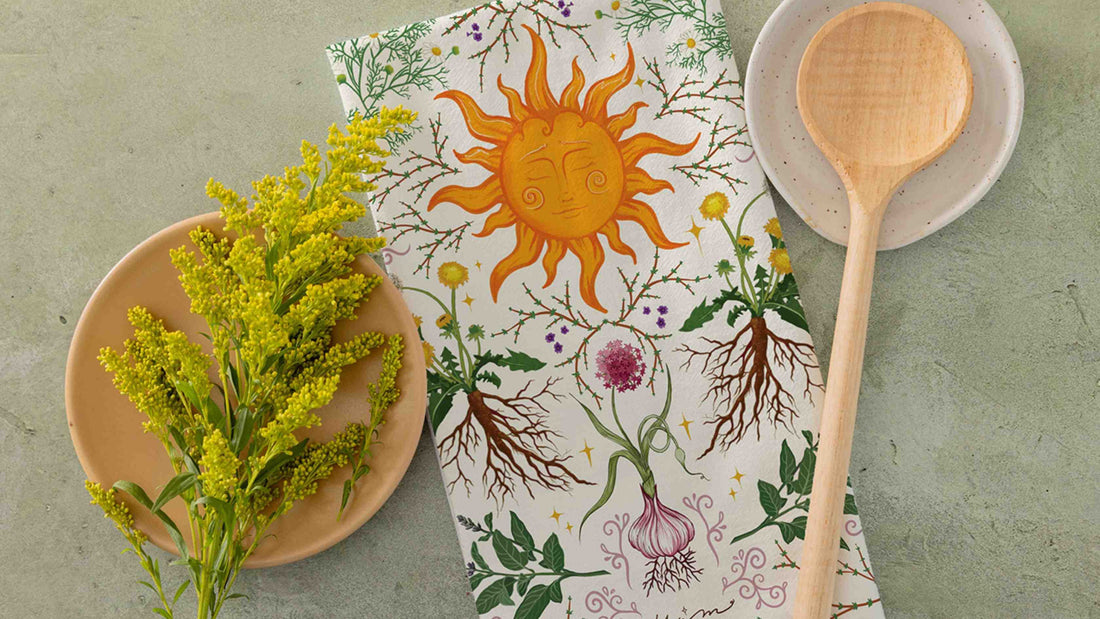 Herbal Kitchen Tea Towel featuring a whimsical botanical illustration with herbs, roots, garlic, wildflowers, and a radiant sun motif, styled alongside fresh yellow flowers and wooden kitchen utensils.