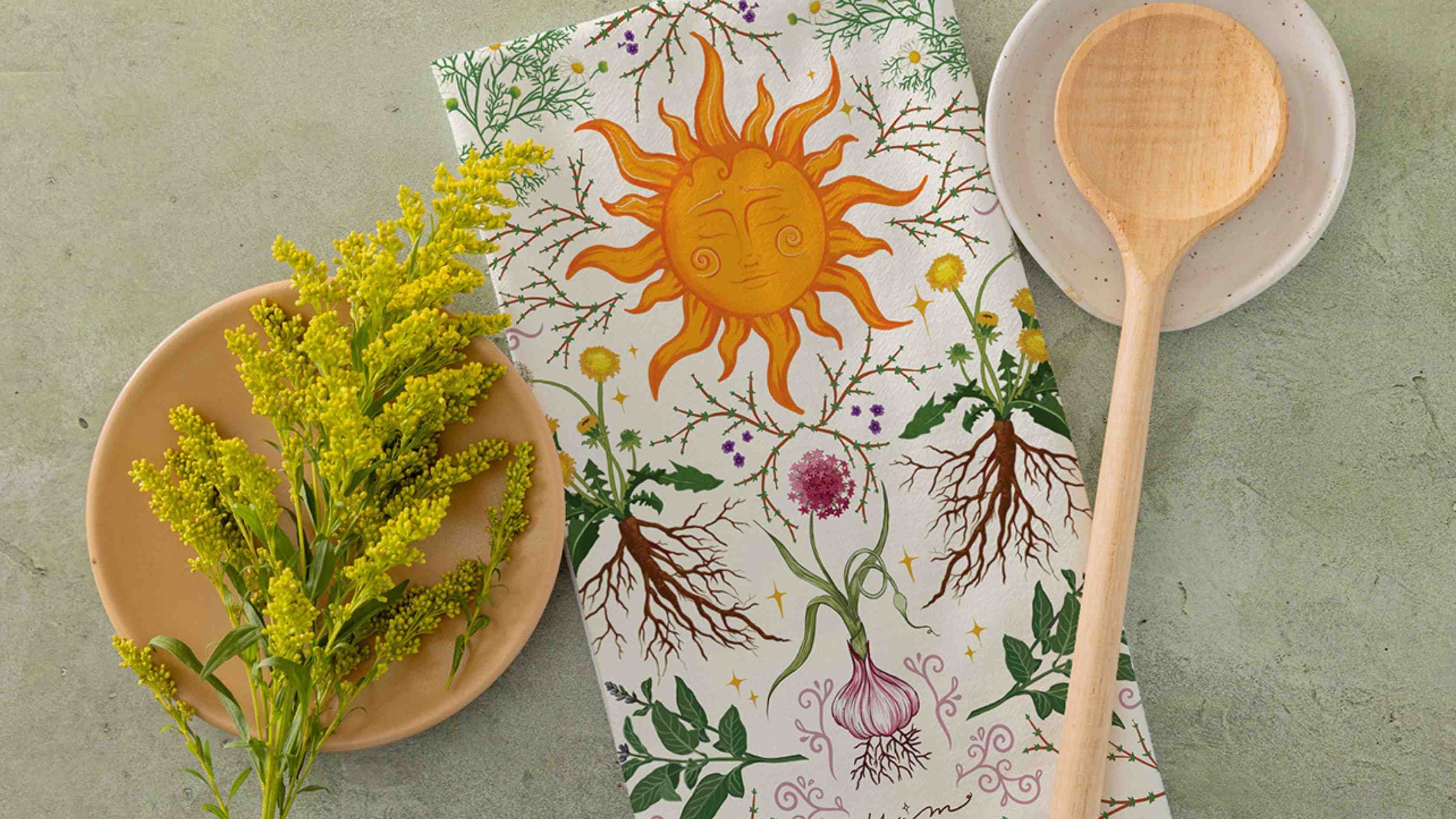 Herbal Kitchen Tea Towel featuring a whimsical botanical illustration with herbs, roots, garlic, wildflowers, and a radiant sun motif, styled alongside fresh yellow flowers and wooden kitchen utensils.