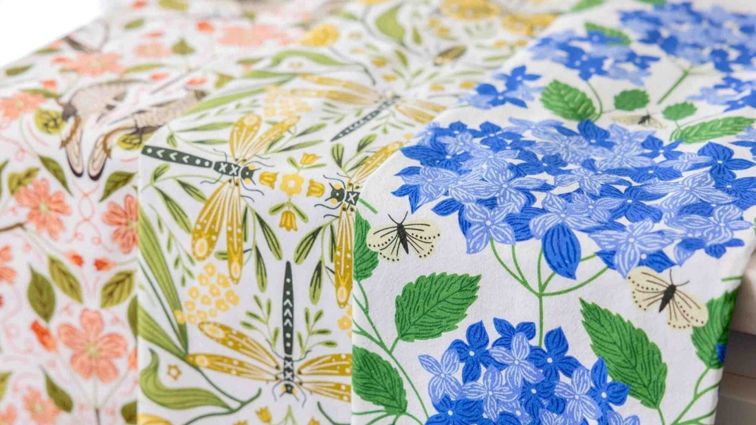 A collection of beautifully patterned Tea Towel Club designs draped side by side, featuring blue hydrangeas, dragonflies, wildflowers, butterflies, and botanical illustrations in vibrant seasonal colors.