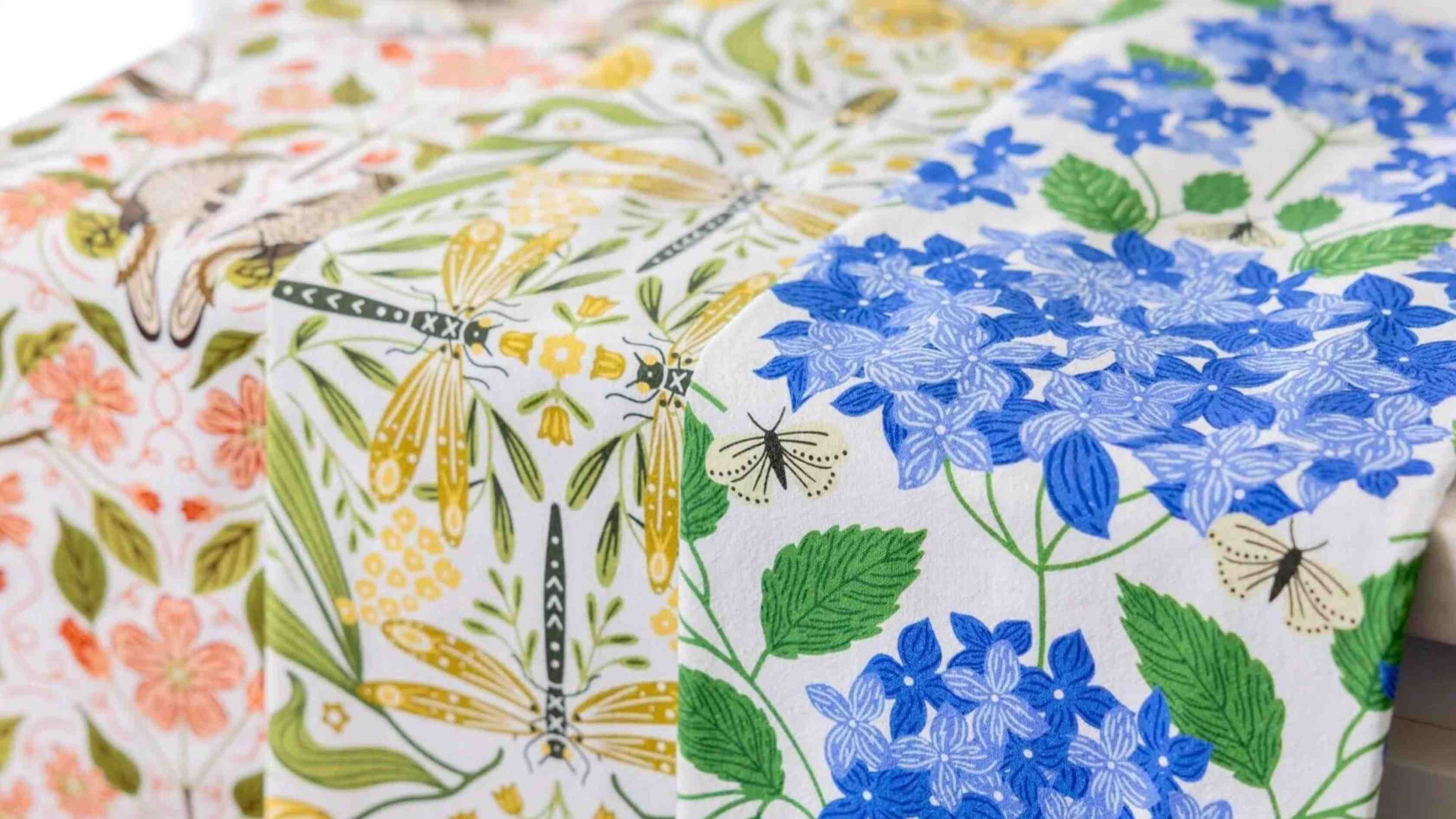 A collection of beautifully patterned Tea Towel Club designs draped side by side, featuring blue hydrangeas, dragonflies, wildflowers, butterflies, and botanical illustrations in vibrant seasonal colors.