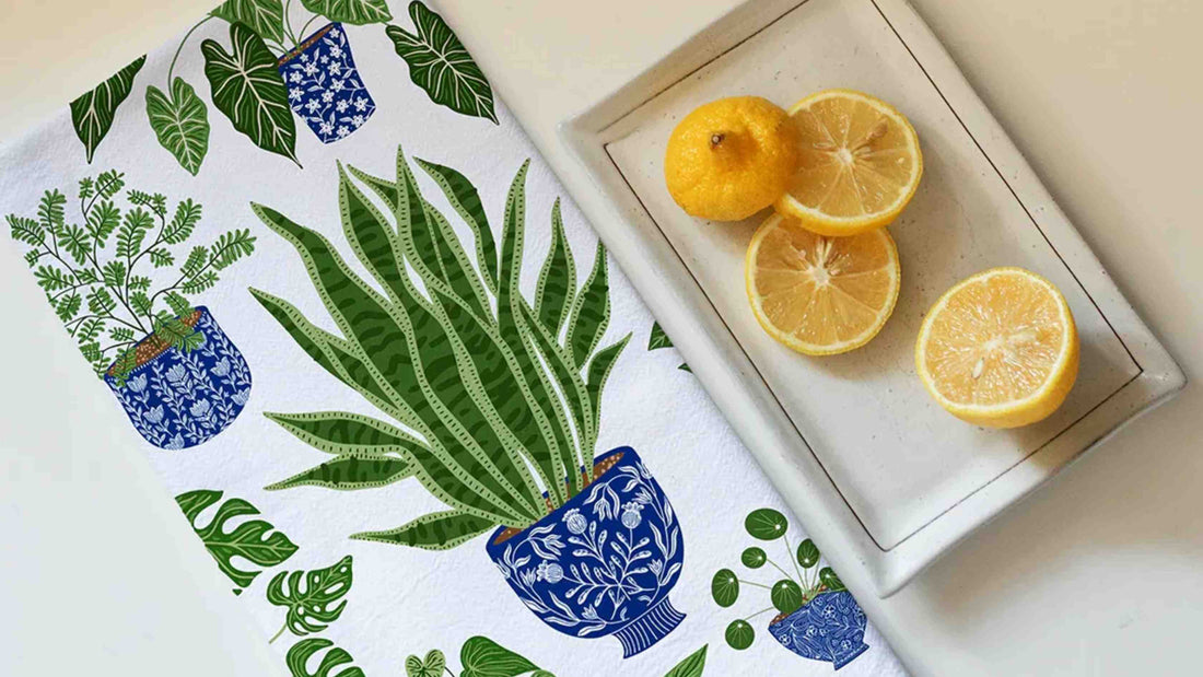 Houseplant Tea Towel featuring illustrated potted houseplants in blue ceramic pots with lush green foliage, styled beside fresh lemons on a tray.