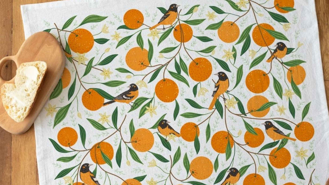 Orange Grove Tea Towel featuring vibrant oranges, leafy branches, and colorful songbirds, styled on a wooden table beside a wooden cutting board with fresh sourdough bread.