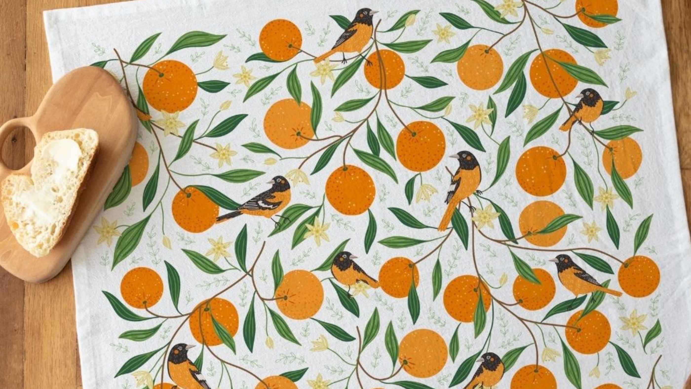Orange Grove Tea Towel featuring vibrant oranges, leafy branches, and colorful songbirds, styled on a wooden table beside a wooden cutting board with fresh sourdough bread.