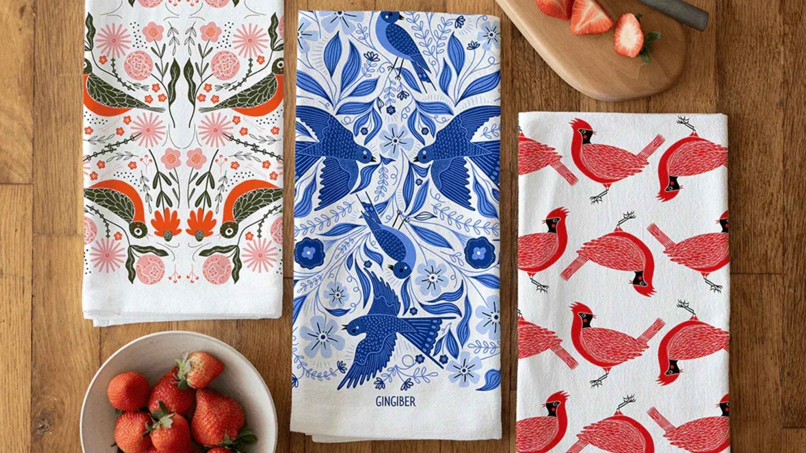 Three Bird Tea Towel Bundle featuring three coordinating cotton tea towels with colorful bird and botanical illustrations, arranged on a wooden table beside fresh strawberries and a cutting board.