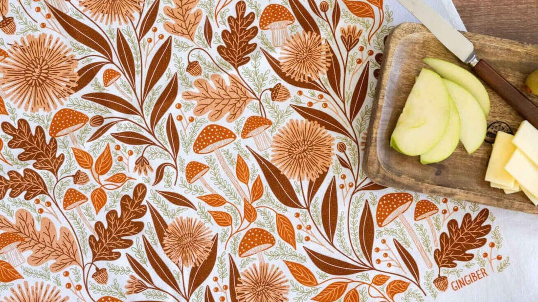 Mushroom Medley Tea Towel featuring autumn-inspired mushrooms, oak leaves, acorns, and botanical foliage in warm earthy tones, styled beside a wooden cutting board with sliced apple and cheese.