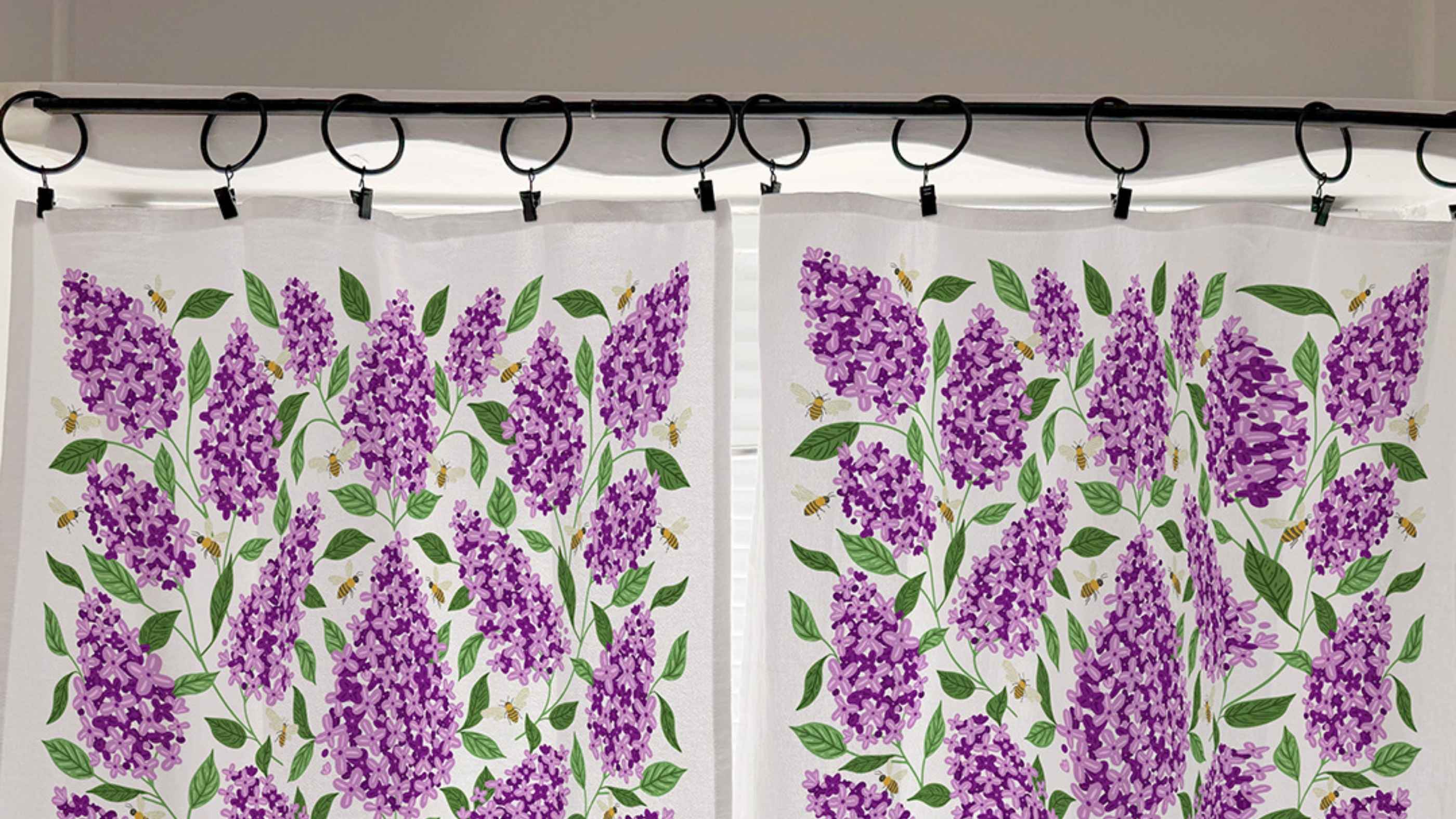 Lilac Café Curtains hanging from black curtain rings, featuring blooming purple lilacs, green leaves, and buzzing bees.