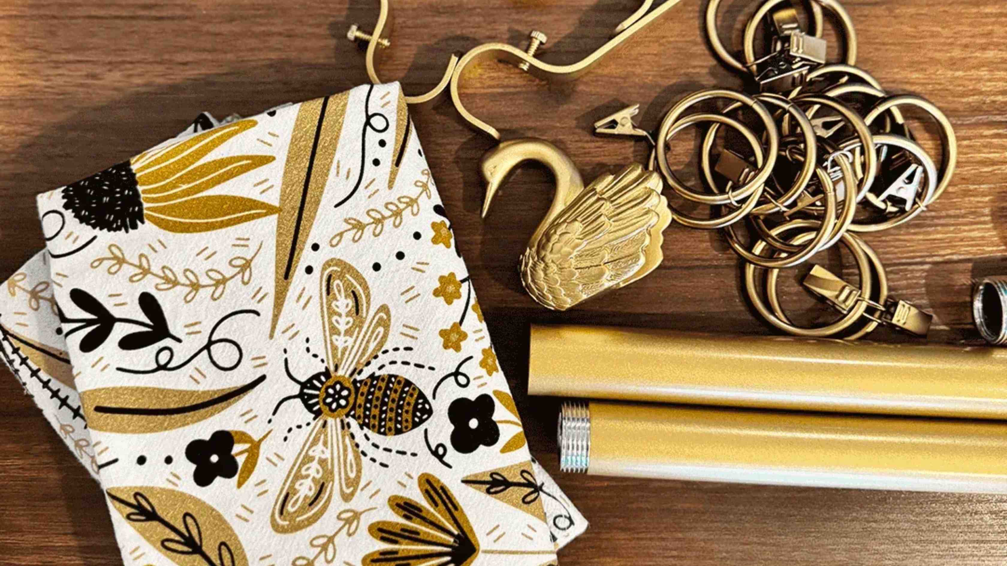 Bee Garden Café Curtains folded beside gold curtain rings and hardware, showcasing a whimsical bee and botanical print in black, white, and metallic gold.