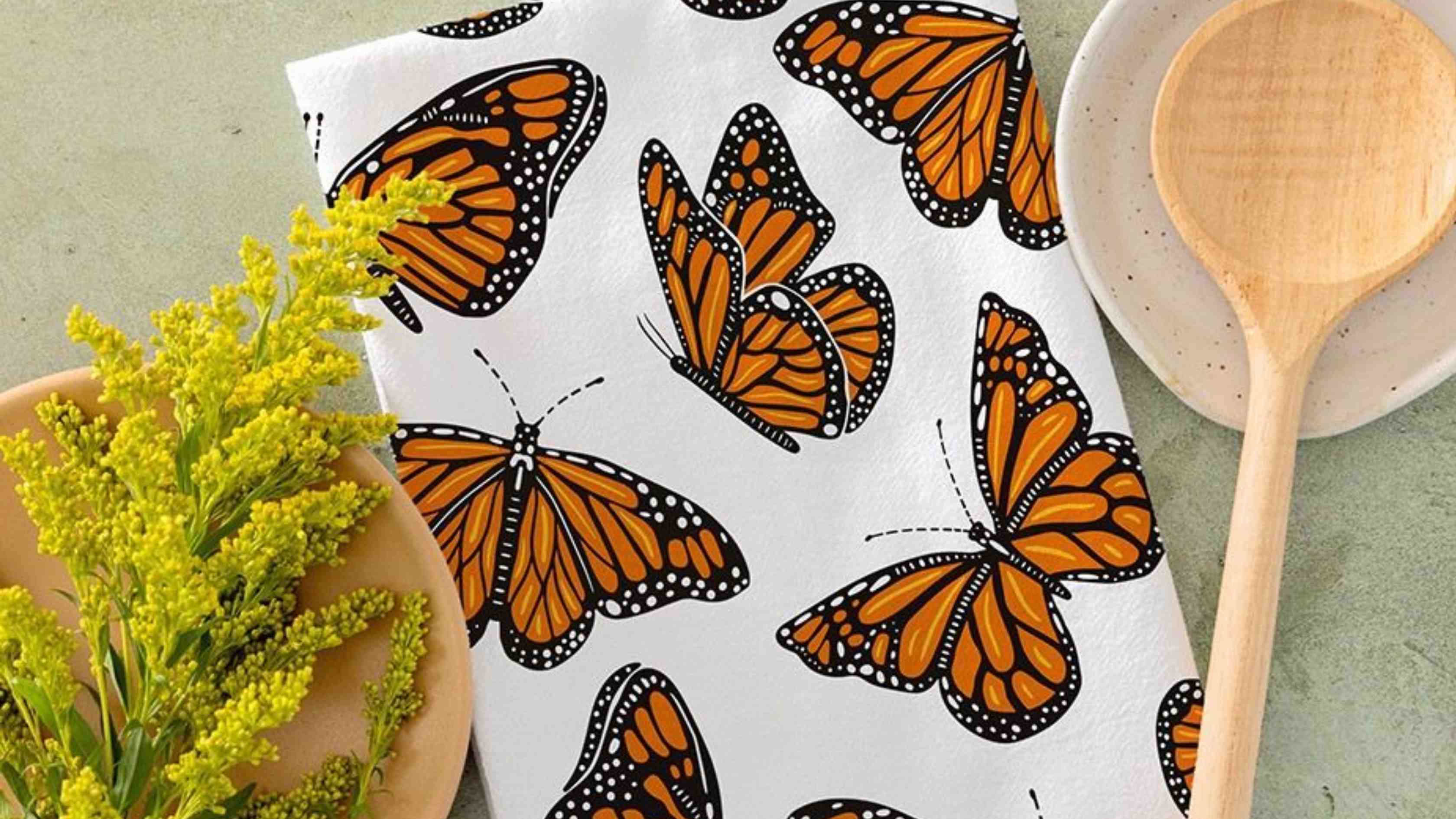 Monarch Flight Tea Towel featuring vibrant orange and black monarch butterflies, styled with fresh yellow flowers and wooden kitchen utensils on a countertop.