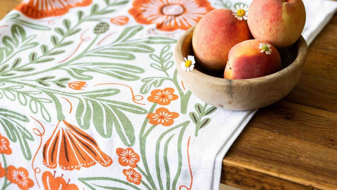 Poppy Tea Towel featuring vibrant orange poppies and soft green botanical foliage, styled beneath a wooden bowl of fresh peaches on a rustic kitchen table.