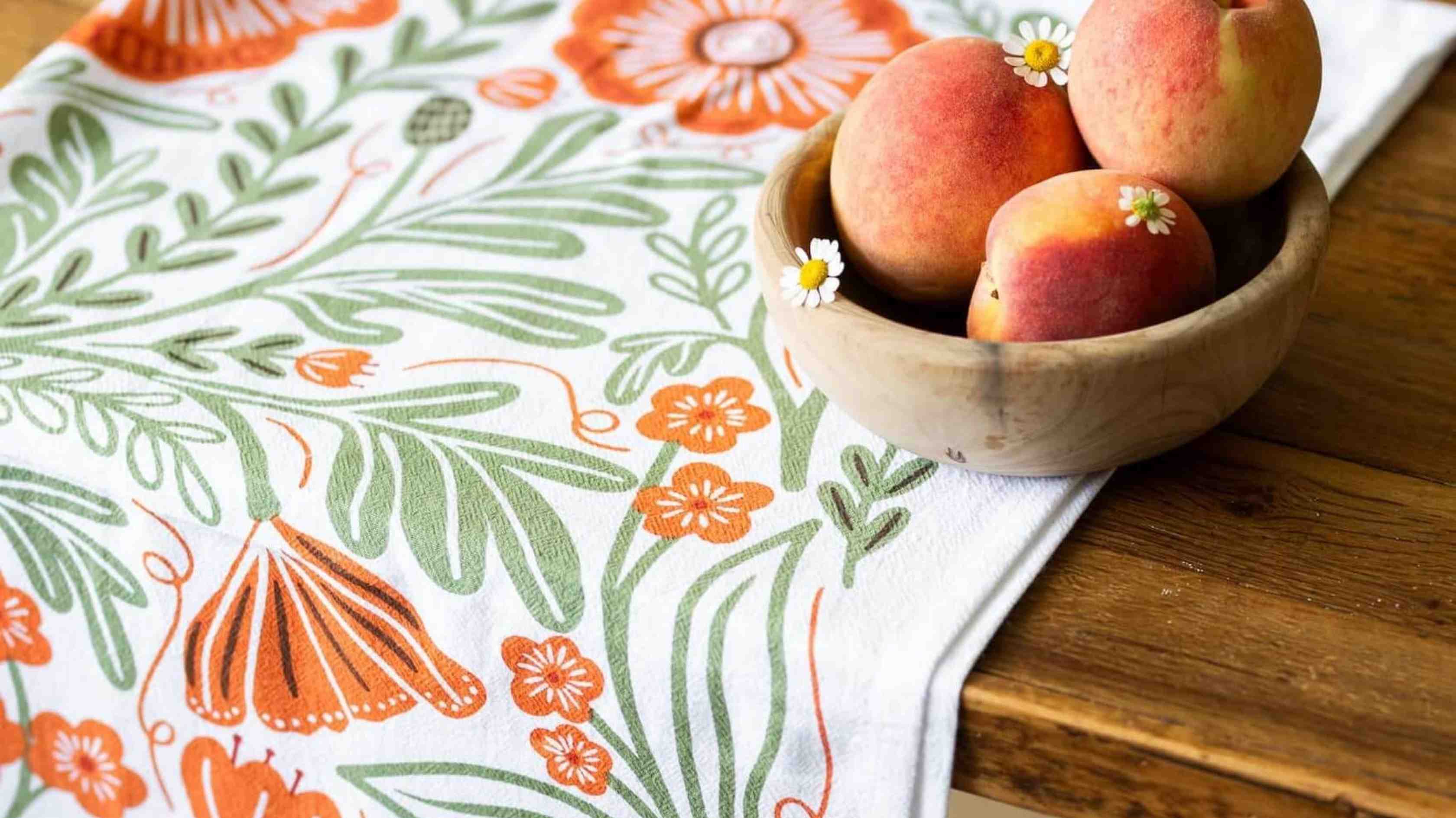 Poppy Tea Towel featuring vibrant orange poppies and soft green botanical foliage, styled beneath a wooden bowl of fresh peaches on a rustic kitchen table.