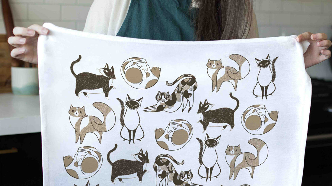 Cat Tea Towel featuring playful illustrated cats in a variety of poses, held up in a bright kitchen to showcase the whimsical black, white, and tan design.