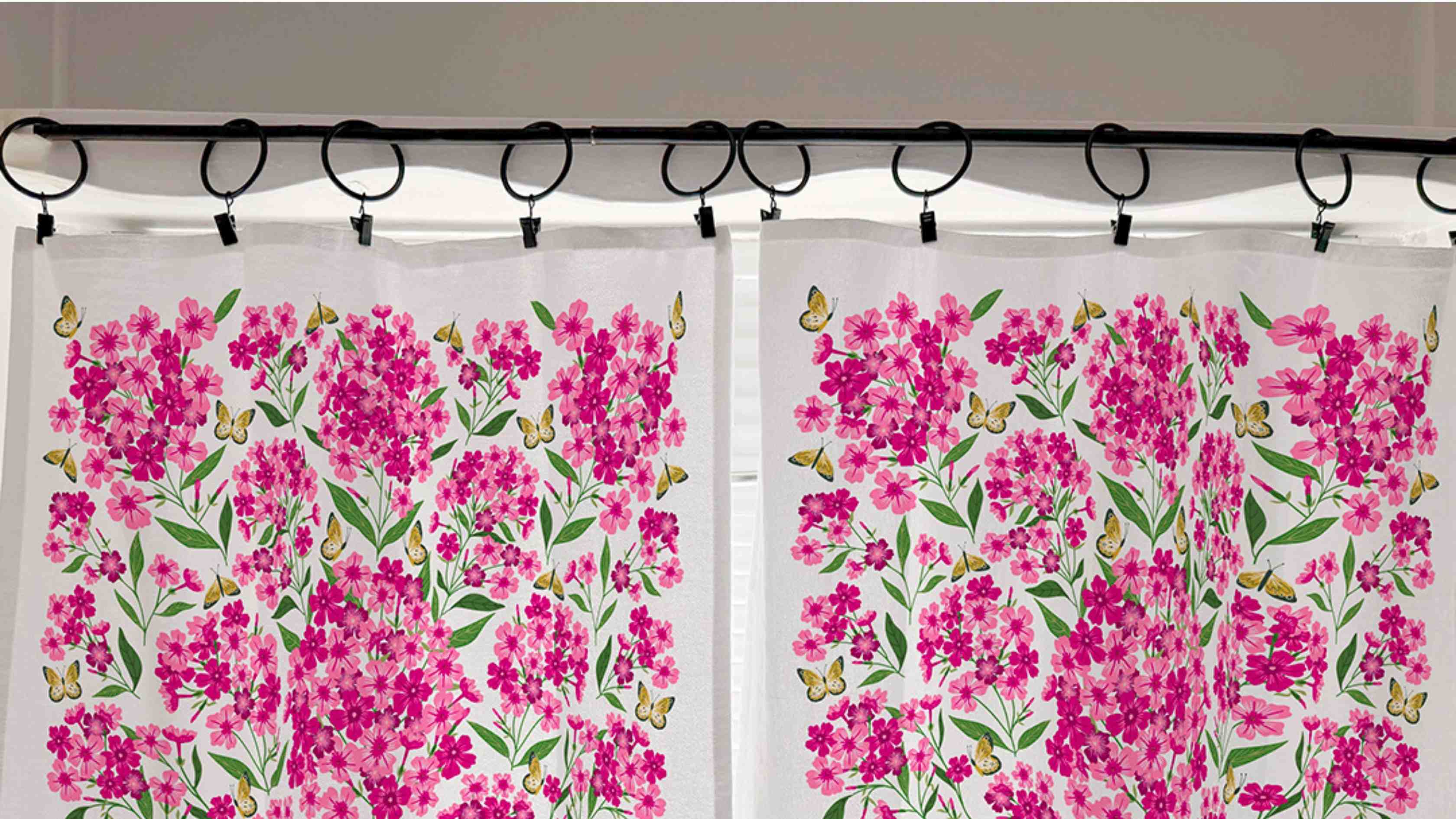 Pink Perennial Café Curtains hanging from black curtain rings, featuring vibrant pink perennial flowers, green foliage, and delicate butterflies.