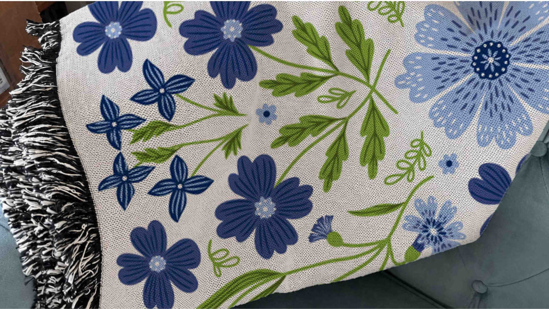 Blue Cornflower Blanket featuring vibrant blue cornflowers and green botanical foliage with soft black and white fringe, draped over a chair.