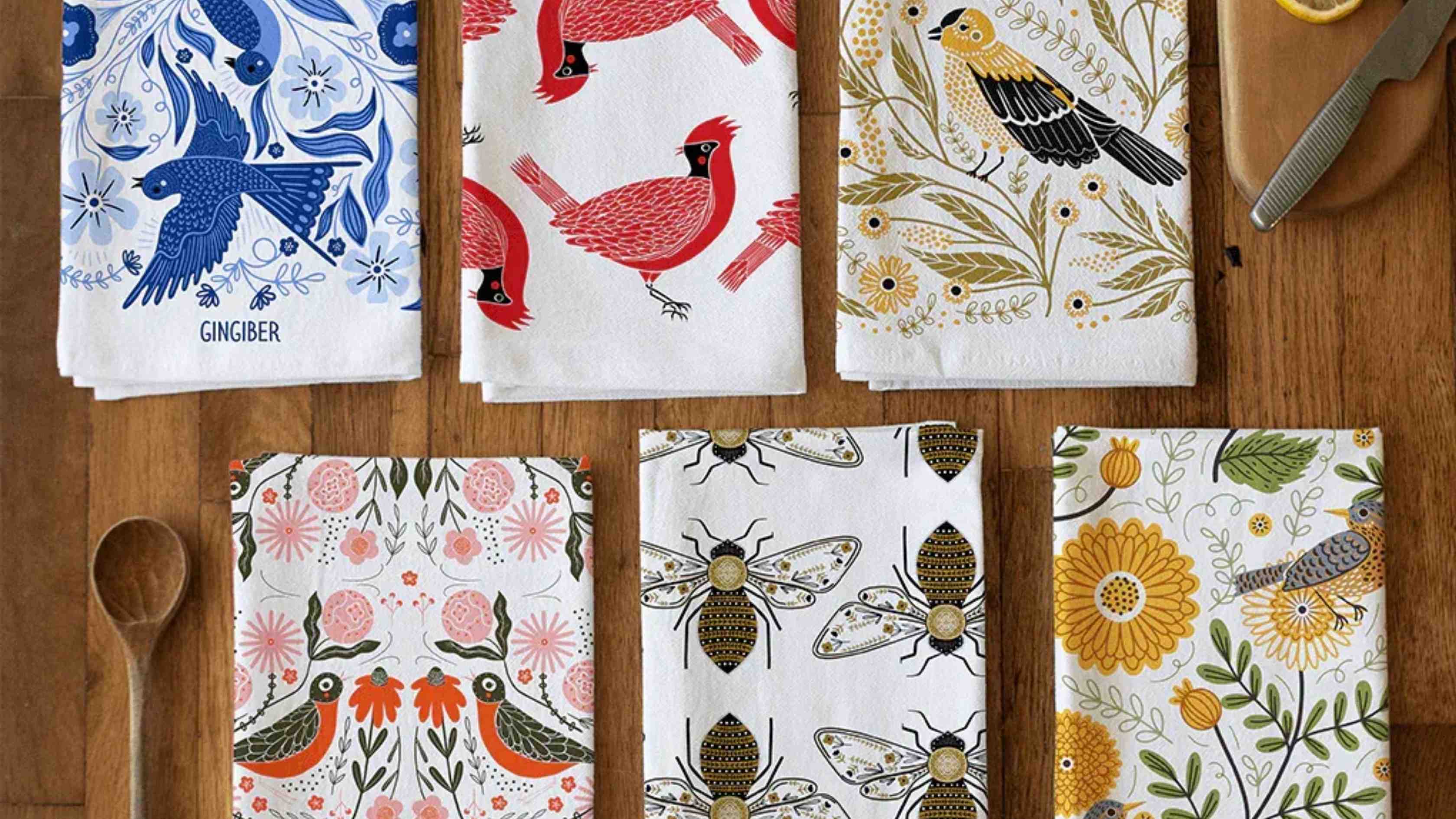Take Flight Bundle featuring six nature-inspired tea towels with colorful bird, bee, and floral illustrations arranged on a wooden tabletop beside wooden kitchen utensils.