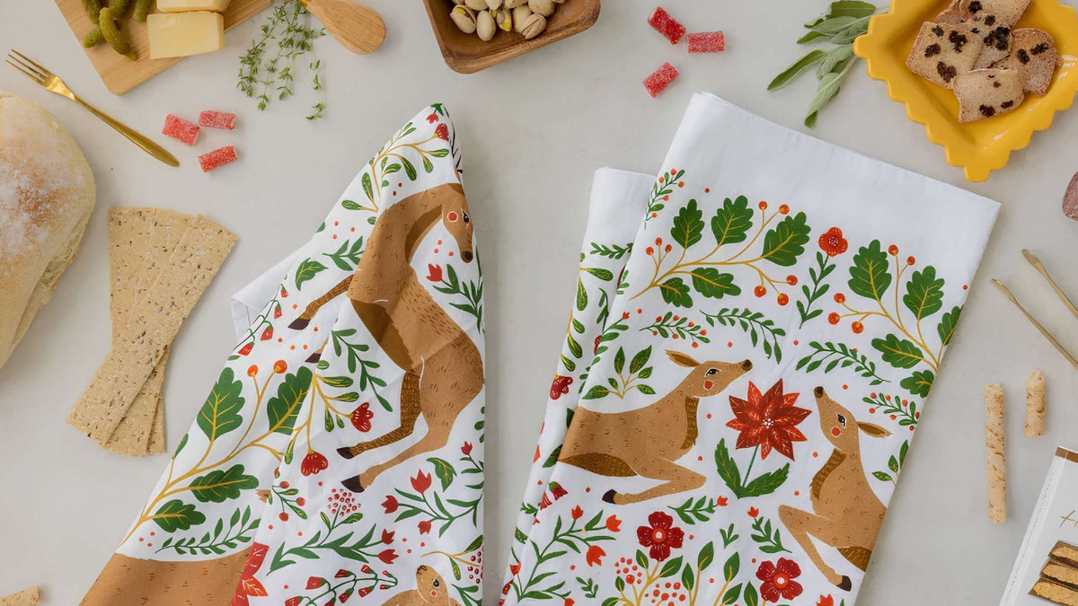 Gingiber Christmas Forest Tea Towels featuring hand-illustrated woodland deer, festive florals, and greenery, styled alongside bread and snacks.