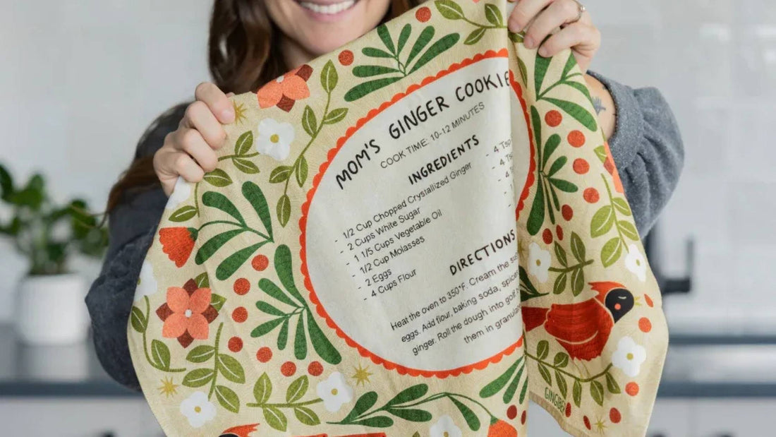 Gingiber Ginger Cookies Recipe Tea Towel featuring Mom's Ginger Cookie recipe, colorful florals, and greenery, held up in a kitchen decorated for holiday baking.