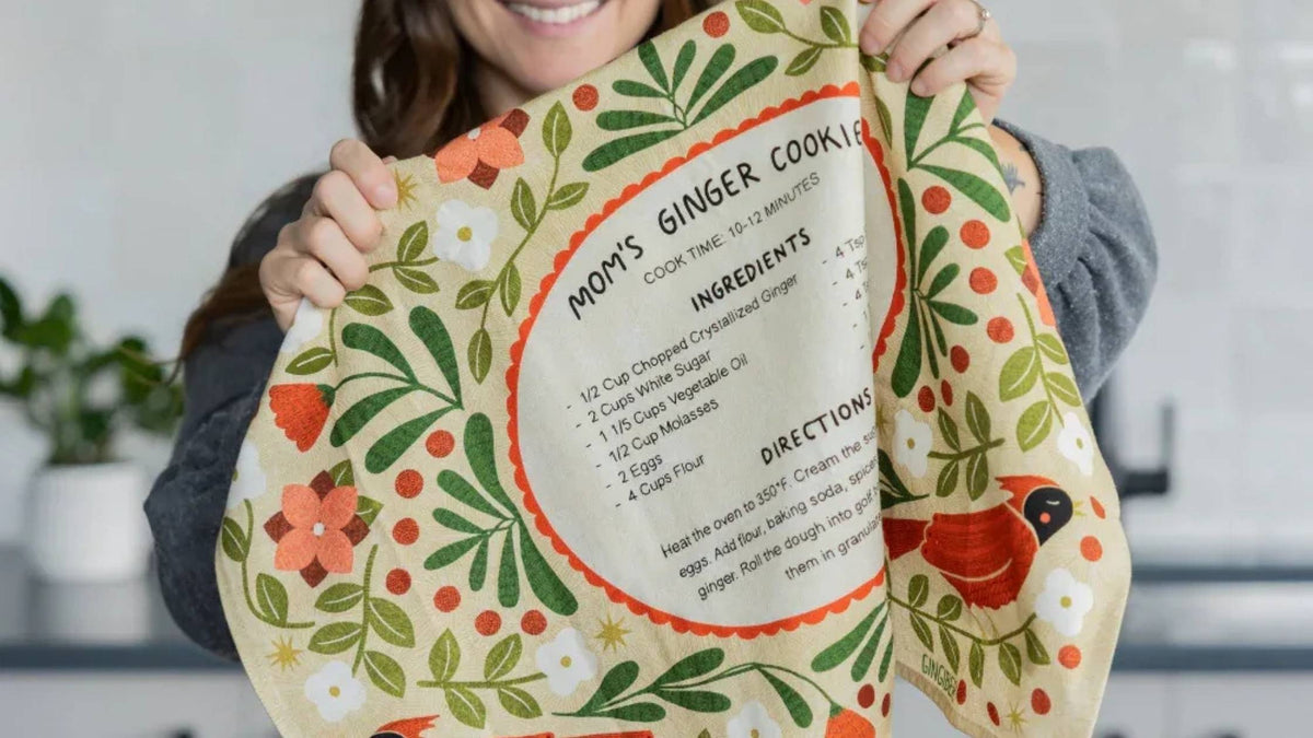 Gingiber Ginger Cookies Recipe Tea Towel featuring Mom's Ginger Cookie recipe, colorful florals, and greenery, held up in a kitchen decorated for holiday baking.