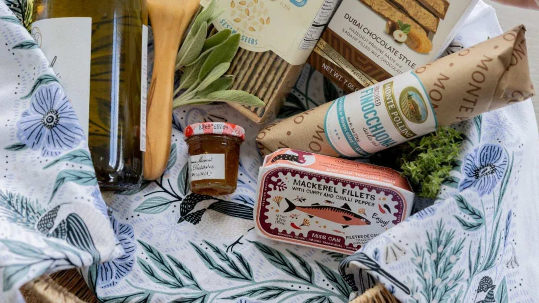 Gingiber Winter Retreat Tea Towel featuring blue floral and bird illustrations, styled in a gift basket with gourmet treats and kitchen essentials.
