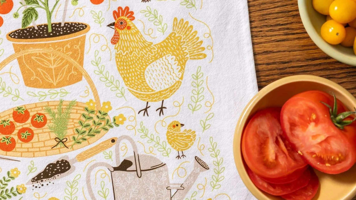 Gingiber Garden Chicken Tea Towel featuring a hand-illustrated hen, chick, garden vegetables, and flowers, styled beside fresh tomatoes on a wooden table.