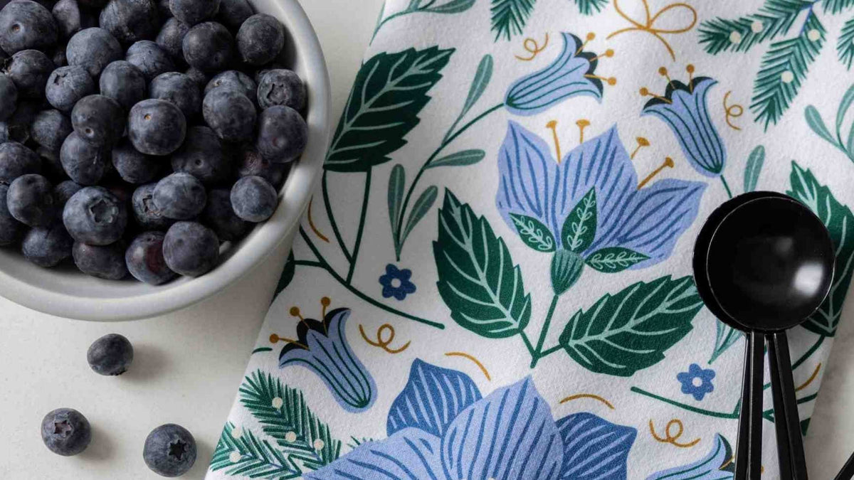 Blue Winter Flowers Tea Towel with blue florals and evergreen details styled beside fresh blueberries and a black measuring spoon.