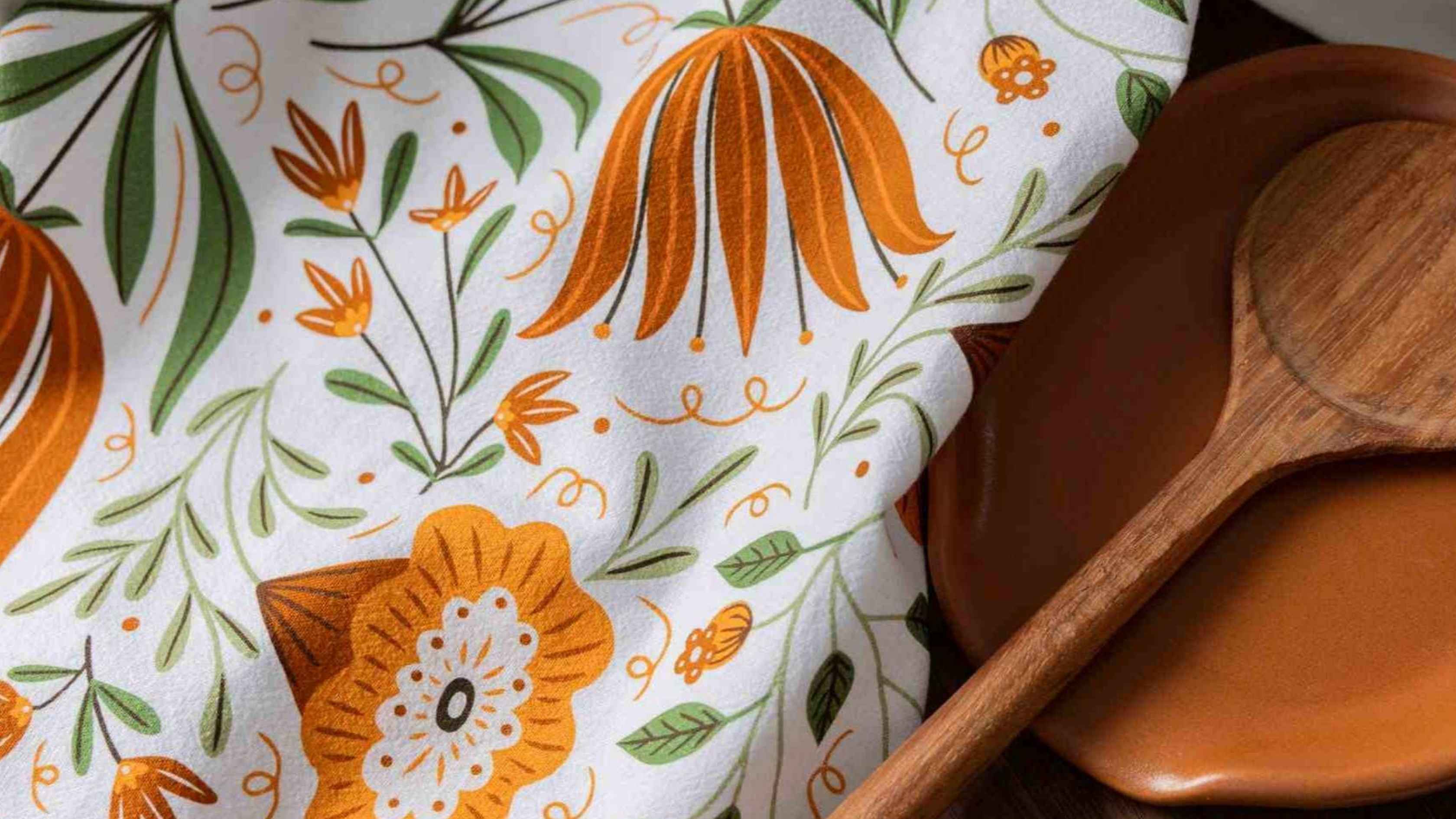 A Folk Floral Tea Towel featuring hand illustrated orange wildflowers and green botanical details on a white background, styled alongside warm wooden bowls and serving utensils.