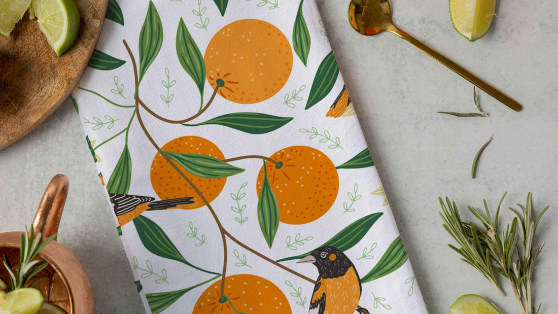 Orange Grove Tea Towel featuring illustrated oranges, green leaves, and colorful birds on a white background, styled with fresh citrus, rosemary, and kitchen accents for a bright summer kitchen look.