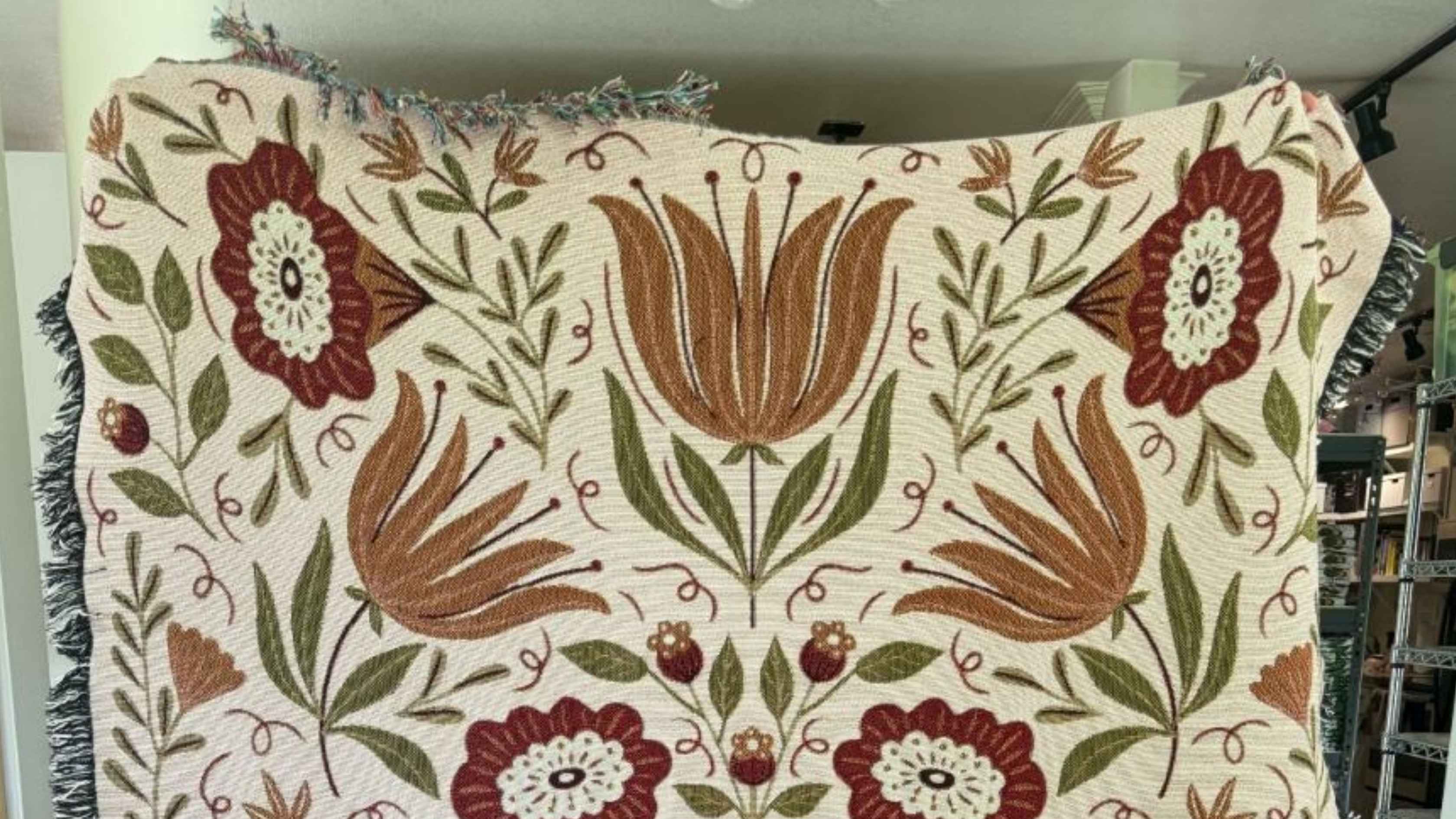 Folk Floral Blanket featuring warm rust, burgundy, and olive green botanical designs on a cream background, with a cozy woven texture and fringed edges, perfect for adding warmth to a coffee lover’s favorite reading or sipping spot.