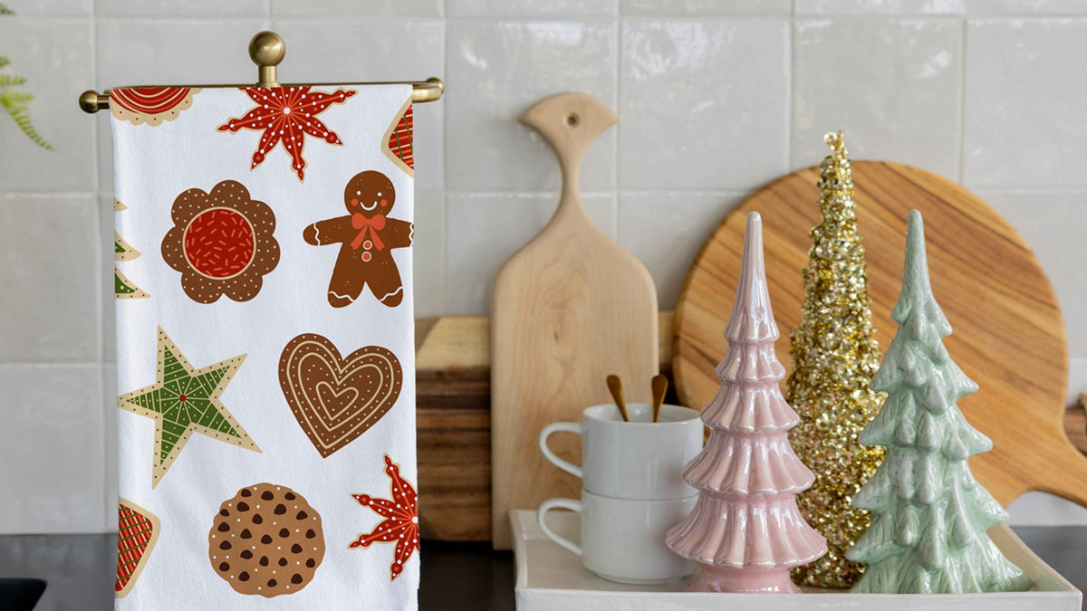 Christmas Cookies Tea Towel featuring festive gingerbread cookies, stars, hearts, and holiday treats, displayed in a cozy kitchen with colorful Christmas tree decorations for a cheerful seasonal touch.