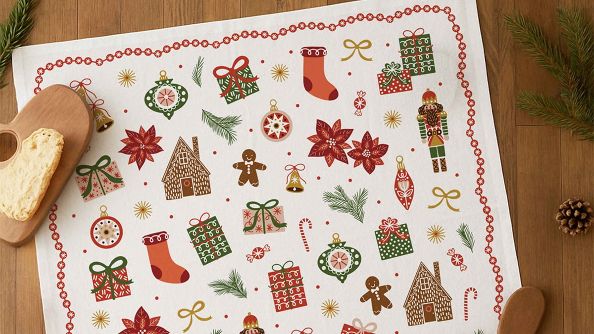 Christmas Tree Tea Towel featuring colorful holiday illustrations of wrapped gifts, ornaments, stockings, gingerbread houses, nutcrackers, poinsettias, and candy canes, styled on a wooden table for a festive white elephant gift idea.
