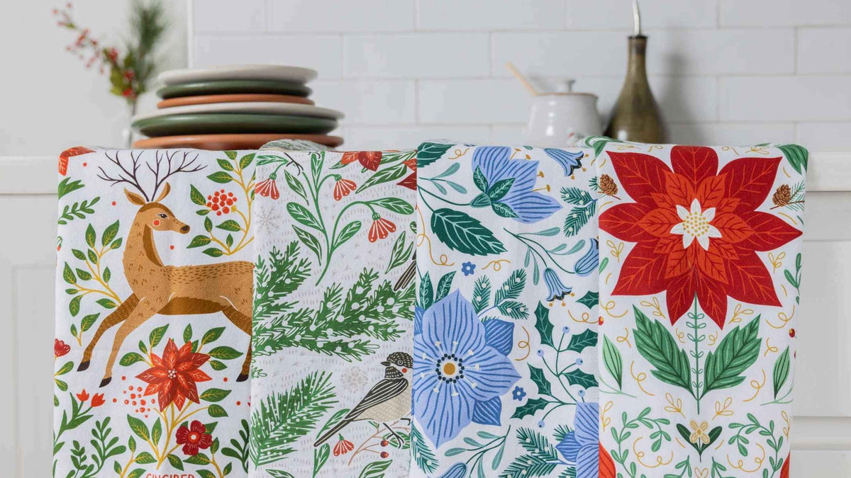 Holiday Best Sellers Tea Towel Bundle featuring festive deer, winter birds, blue florals, greenery, and poinsettia designs displayed in a bright kitchen.