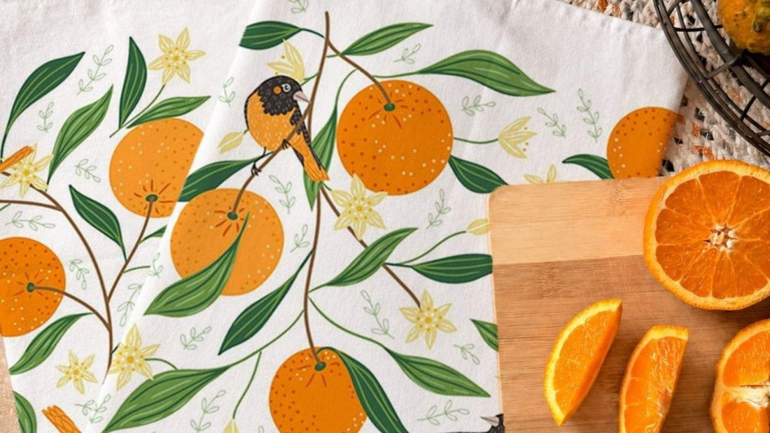 An Orange Grove Tea Towel featuring hand illustrated oranges, leafy branches, blossoms, and a songbird, styled beside fresh orange slices on a cutting board.