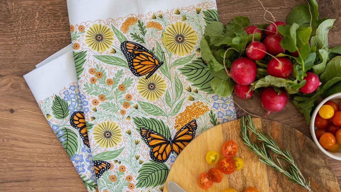 A Monarch Garden Tea Towel featuring hand illustrated monarch butterflies, wildflowers, and leafy botanicals, styled with fresh radishes, cherry tomatoes, and herbs on a rustic wooden table.