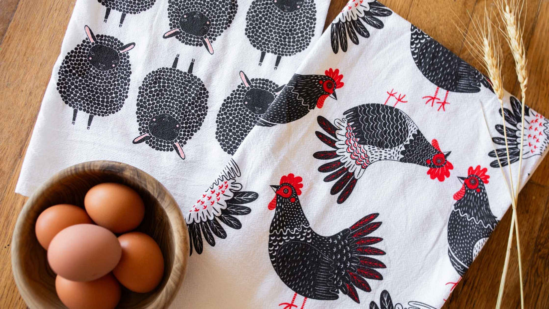 Farm Friends Tea Towel Bundle featuring coordinating sheep and chicken tea towels arranged beside fresh eggs in a wooden bowl and dried wheat on a rustic wooden table. C