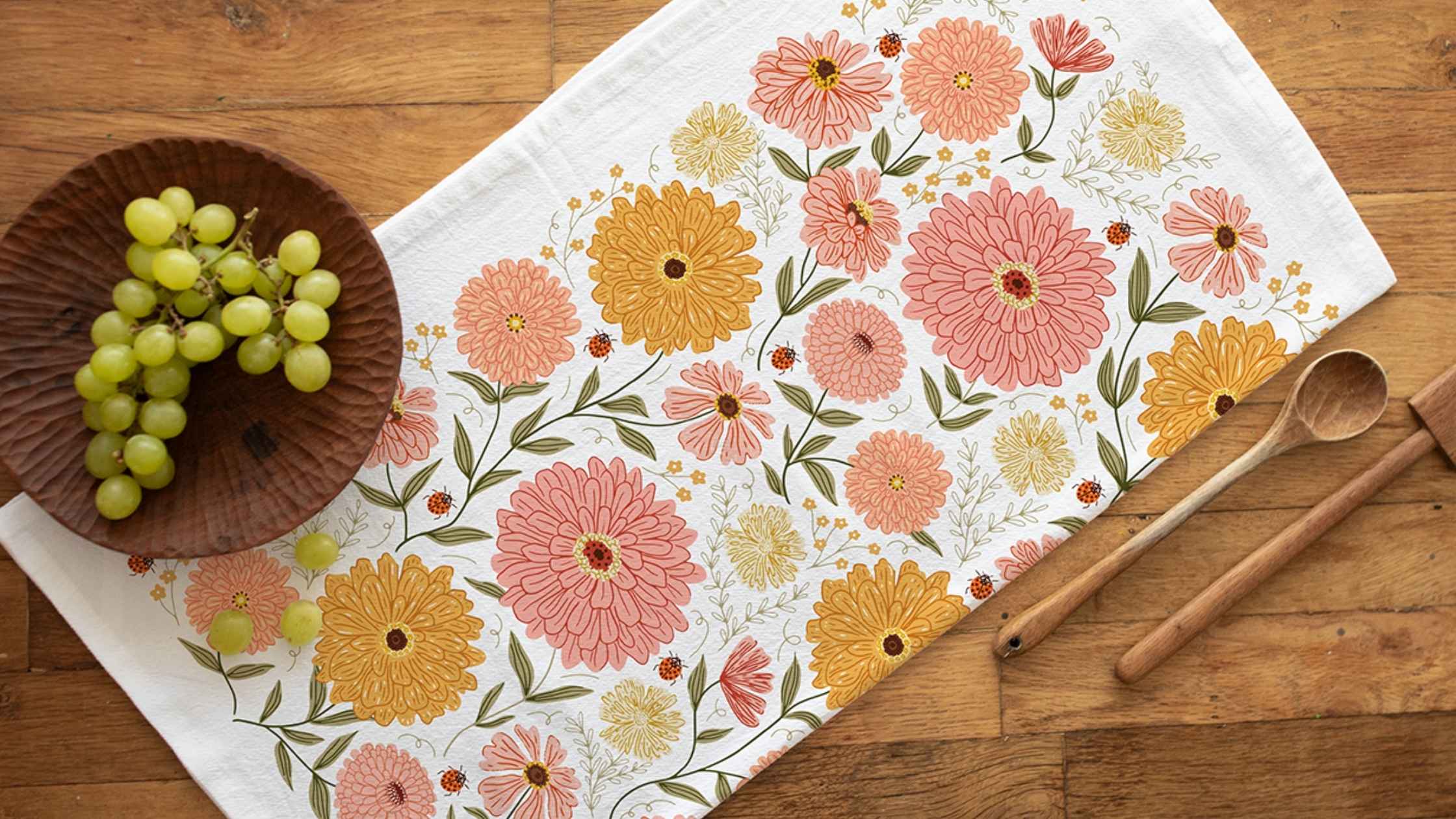 Zinnia Tea Towel featuring cheerful pink, peach, and golden zinnia flowers with delicate greenery, styled beside a wooden bowl of fresh grapes and wooden serving utensils on a rustic table.