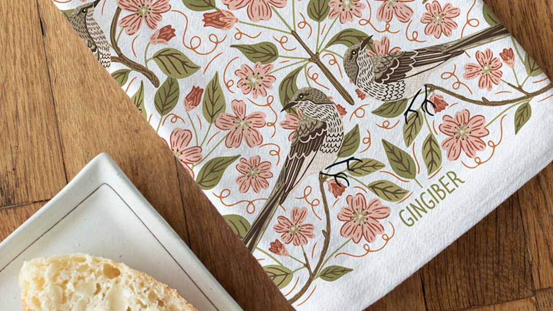 Apple Blossom Tea Towel featuring delicate apple blossoms, leafy branches, and songbirds in soft blush pink, sage green, and warm brown tones, styled beside fresh bread on a rustic wooden table.