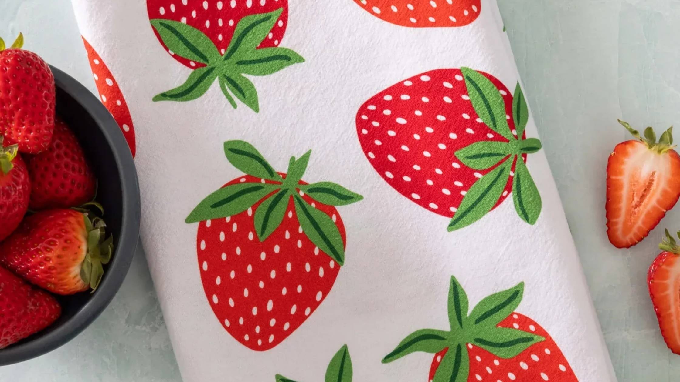 Strawberry Tea Towel featuring bright red strawberries with vibrant green leaves, styled beside a bowl of fresh strawberries and sliced berries on a light countertop.