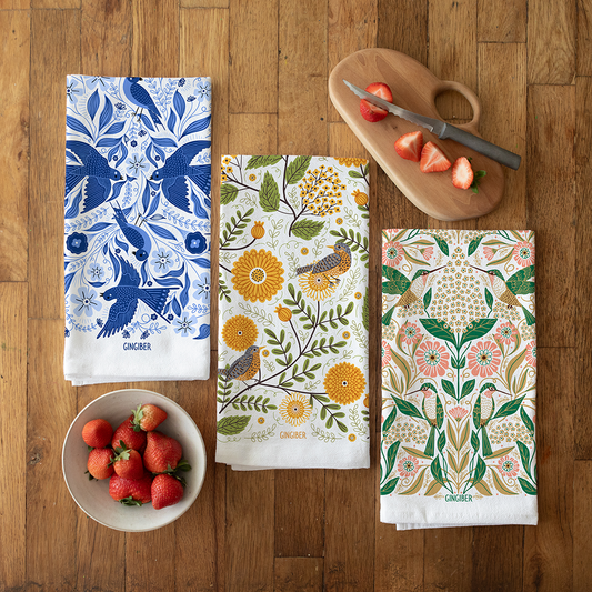 A collection of three bird focused tea towels, folded and displayed on a wooded counter top.