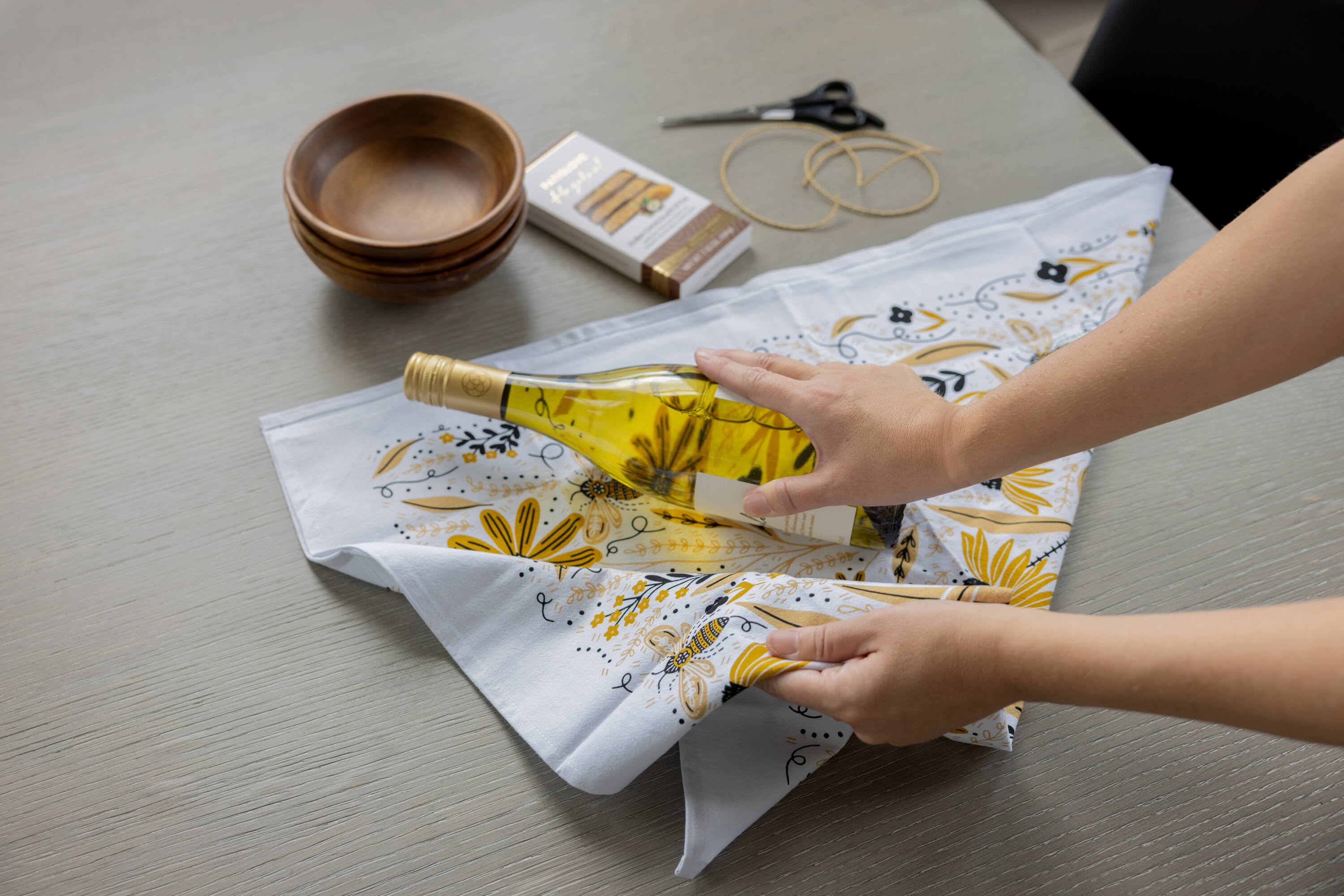 Person wrapping a bottle of olive oil in floral paper on a table with a wooden bowl and scissors.