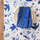 Blue patterned napkin on a white plate with a matching tablecloth featuring blue birds and flowers on a wooden surface.