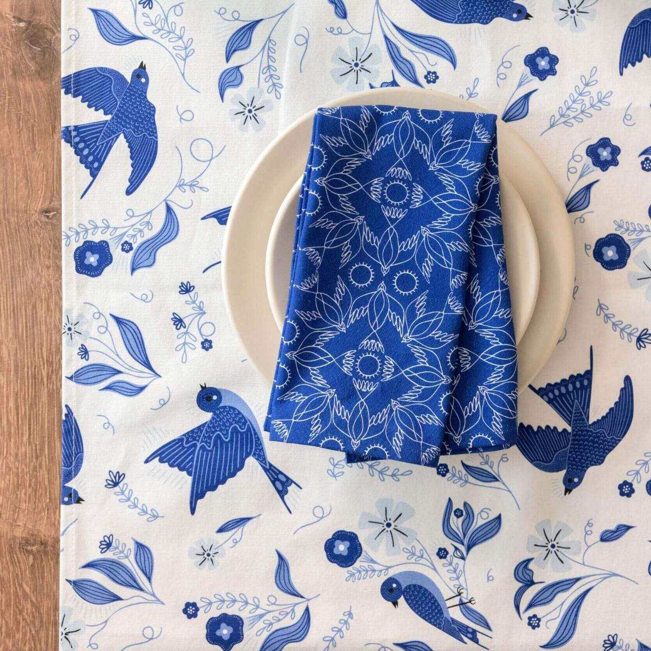 Blue patterned napkin on a white plate with a matching tablecloth featuring blue birds and flowers on a wooden surface.