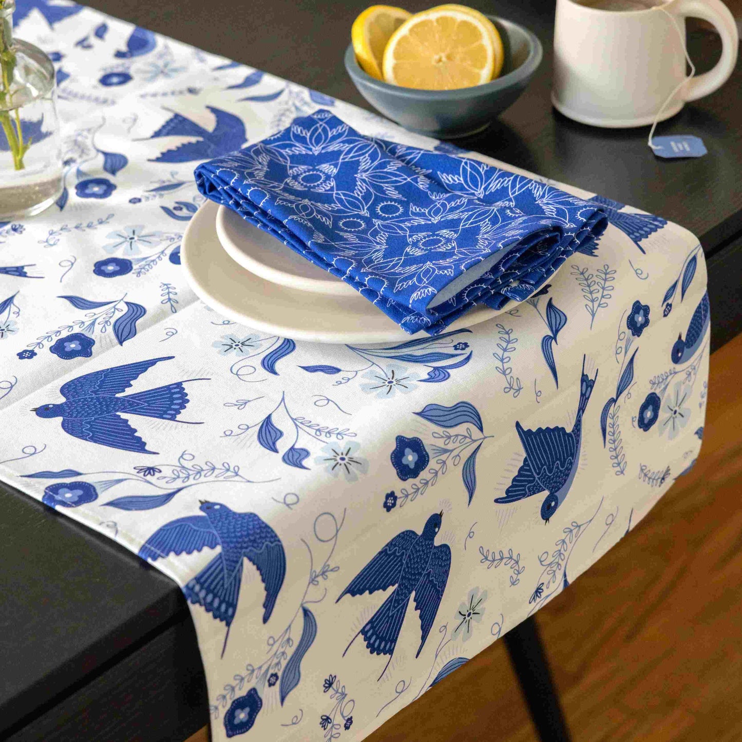 Table setting with a blue and white tablecloth featuring bird patterns, a folded blue napkin, and a bowl of lemons.