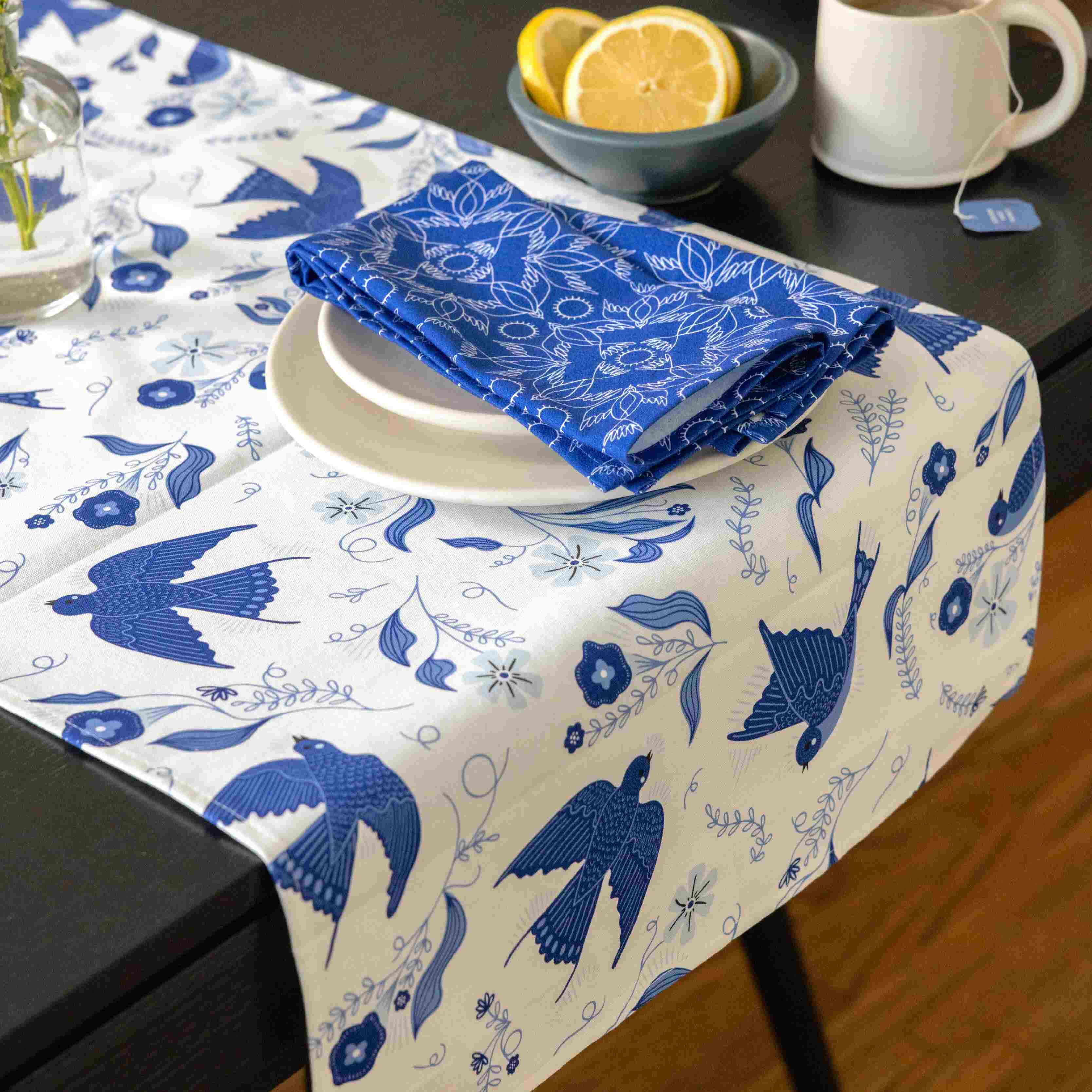 Table setting with a blue and white tablecloth featuring bird patterns, a folded blue napkin, and a bowl of lemons.