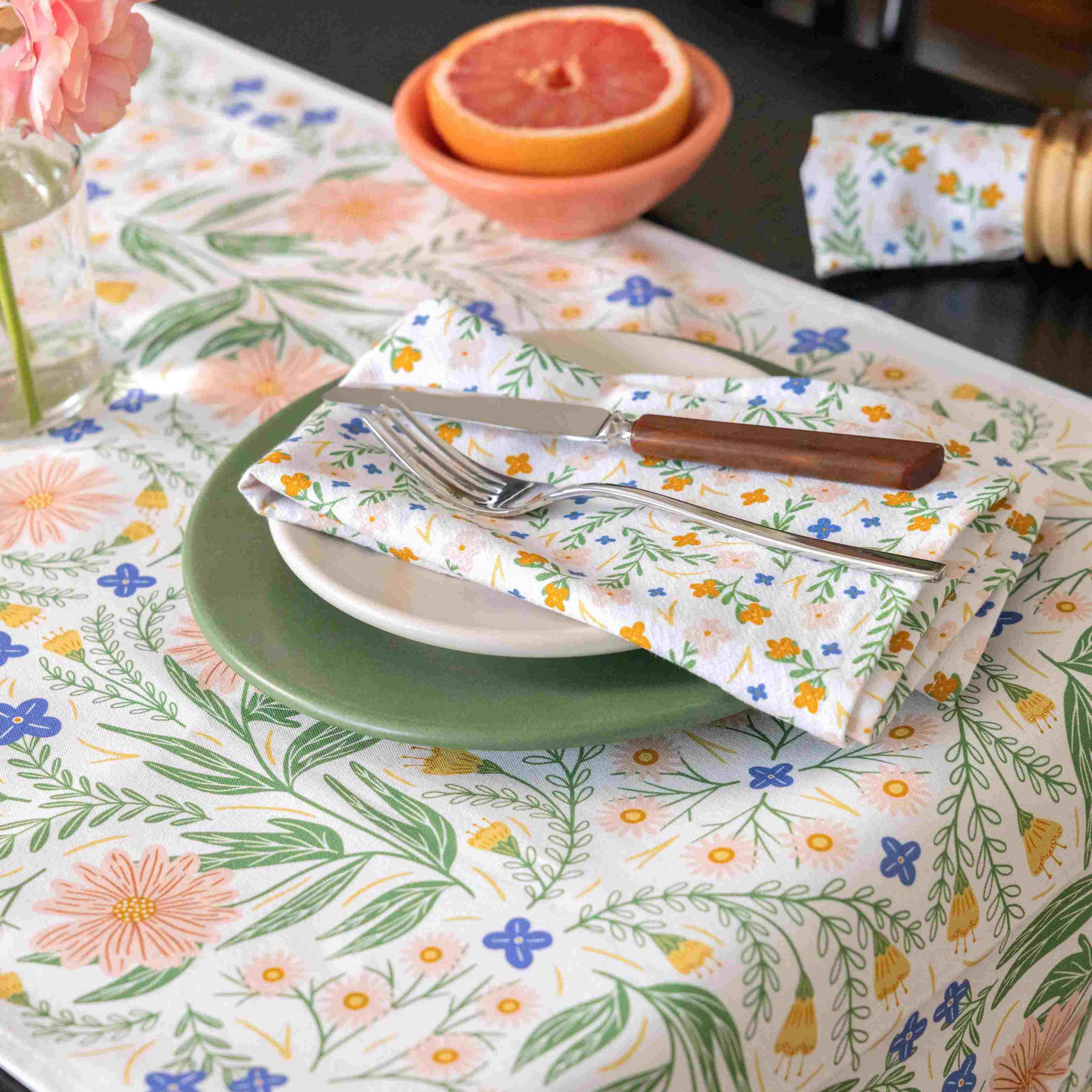 Table setting with floral tablecloth, plates, cutlery, and a bowl of fruit.