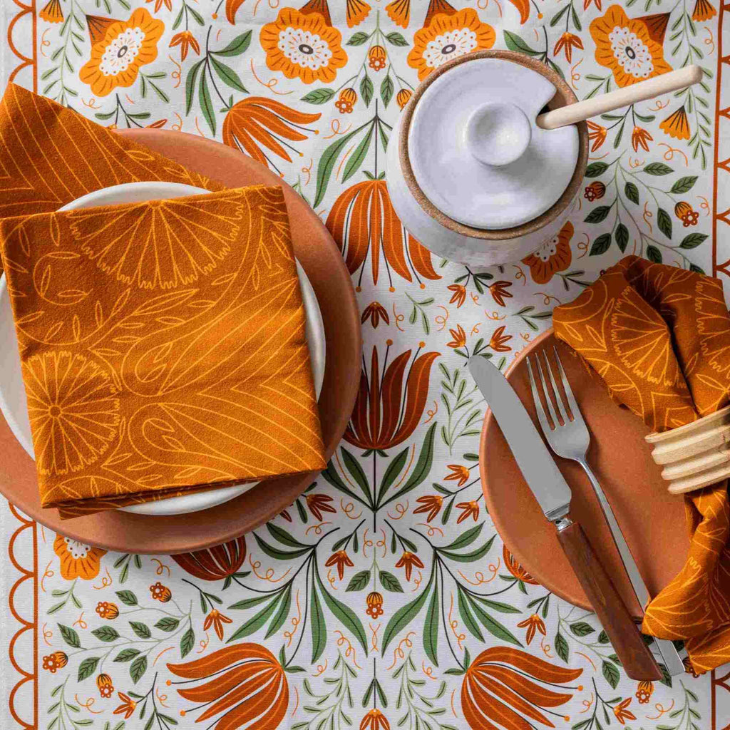 A styled tabletop setting featuring a white tablecloth with a bold orange and green floral tulip pattern, orange plates, matching napkins with a sunburst print, a white lidded container with a wooden spoon, and cutlery.