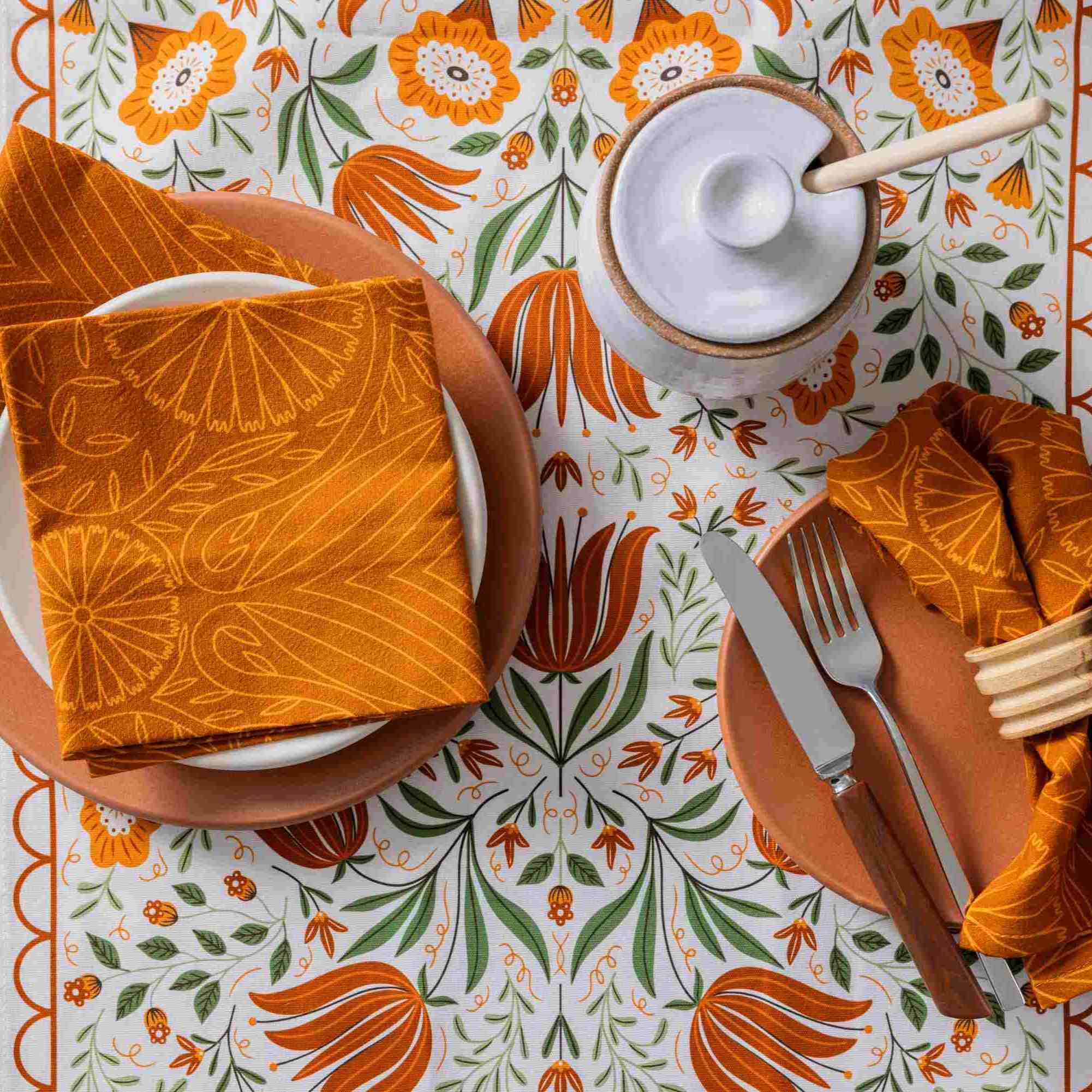 A styled tabletop setting featuring a white tablecloth with a bold orange and green floral tulip pattern, orange plates, matching napkins with a sunburst print, a white lidded container with a wooden spoon, and cutlery.