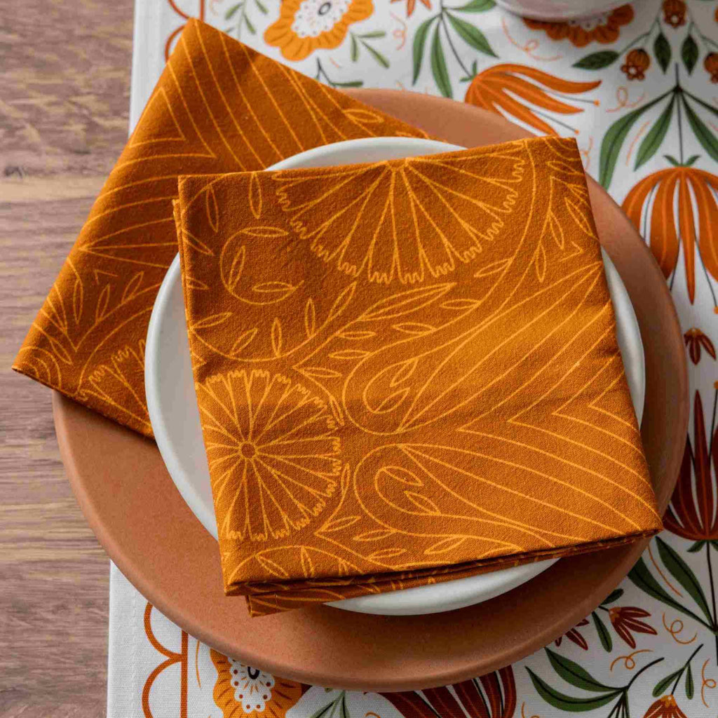 An orange patterned fabric napkin with a yellow-orange floral and leaf design is folded over a white plate on a brown ceramic plate, set on a matching patterned table runner.